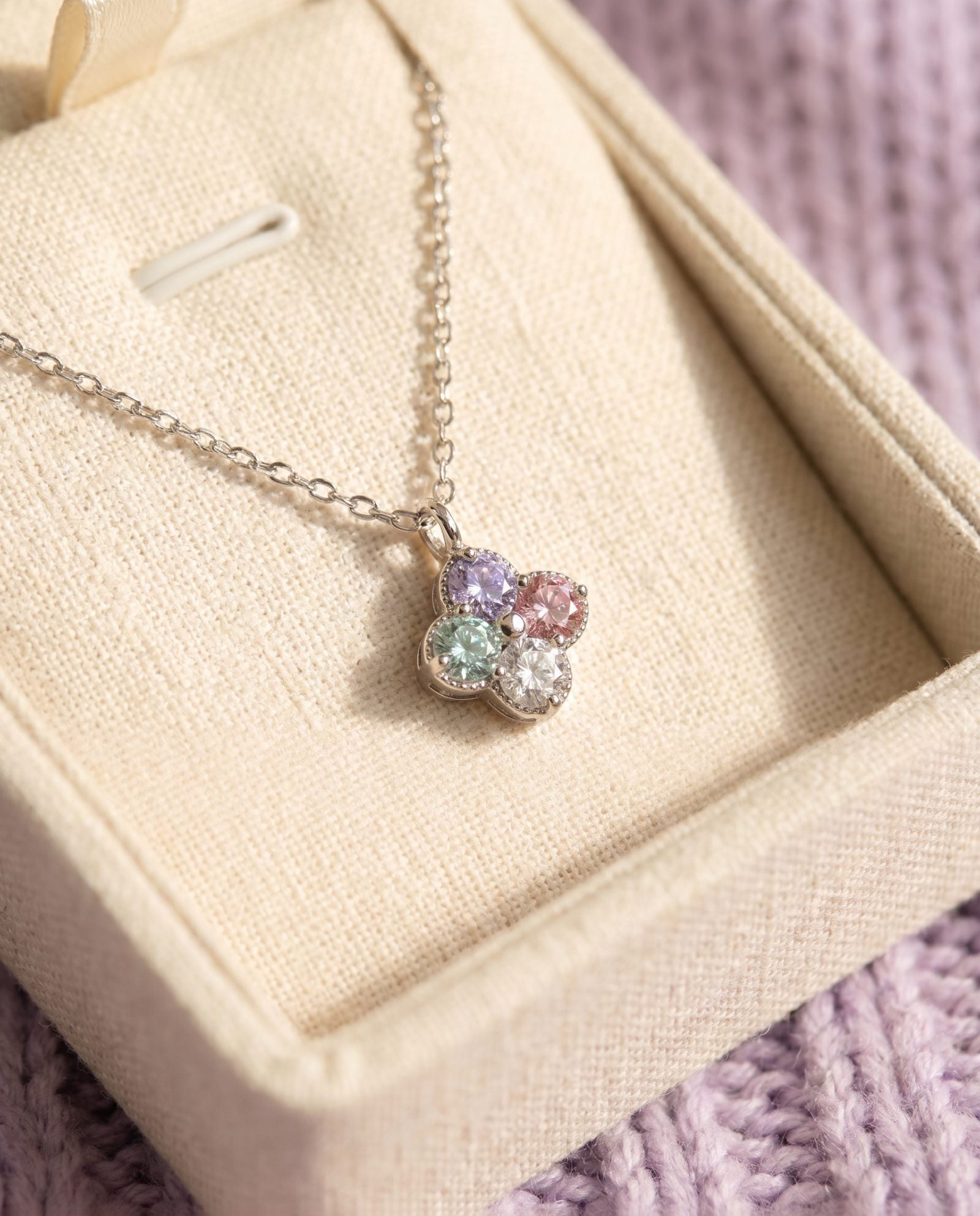 The A Tale of Two Clovers necklace resting in a premium cream jewelry box against a soft lavender knit background.