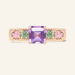A rose gold ring featuring a central square-cut amethyst stone, flanked by two pink tourmalines and two green moissanites on either side, set in a textured band,