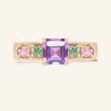 A rose gold ring featuring a central square-cut amethyst stone, flanked by two pink tourmalines and two green moissanites on either side, set in a textured band,