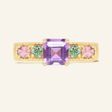 A yellow gold ring featuring a central square-cut amethyst stone, flanked by two pink tourmalines and two green moissanites on either side, set in a textured band,