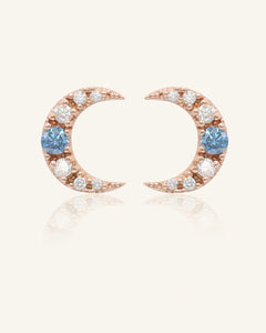 Close-up of the Blue Moissanite Moon Studs in rose gold, resting on a warm linen background