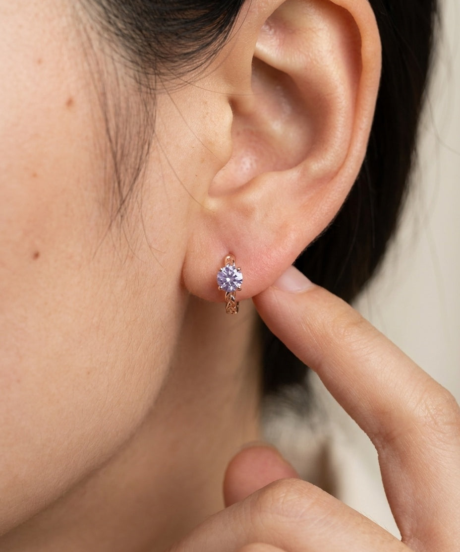 Side profile shot of a model wearing a rose gold vermeil Braided Lavender Moissanite Huggie Hoop (1ctw) with a finger touching the ear.