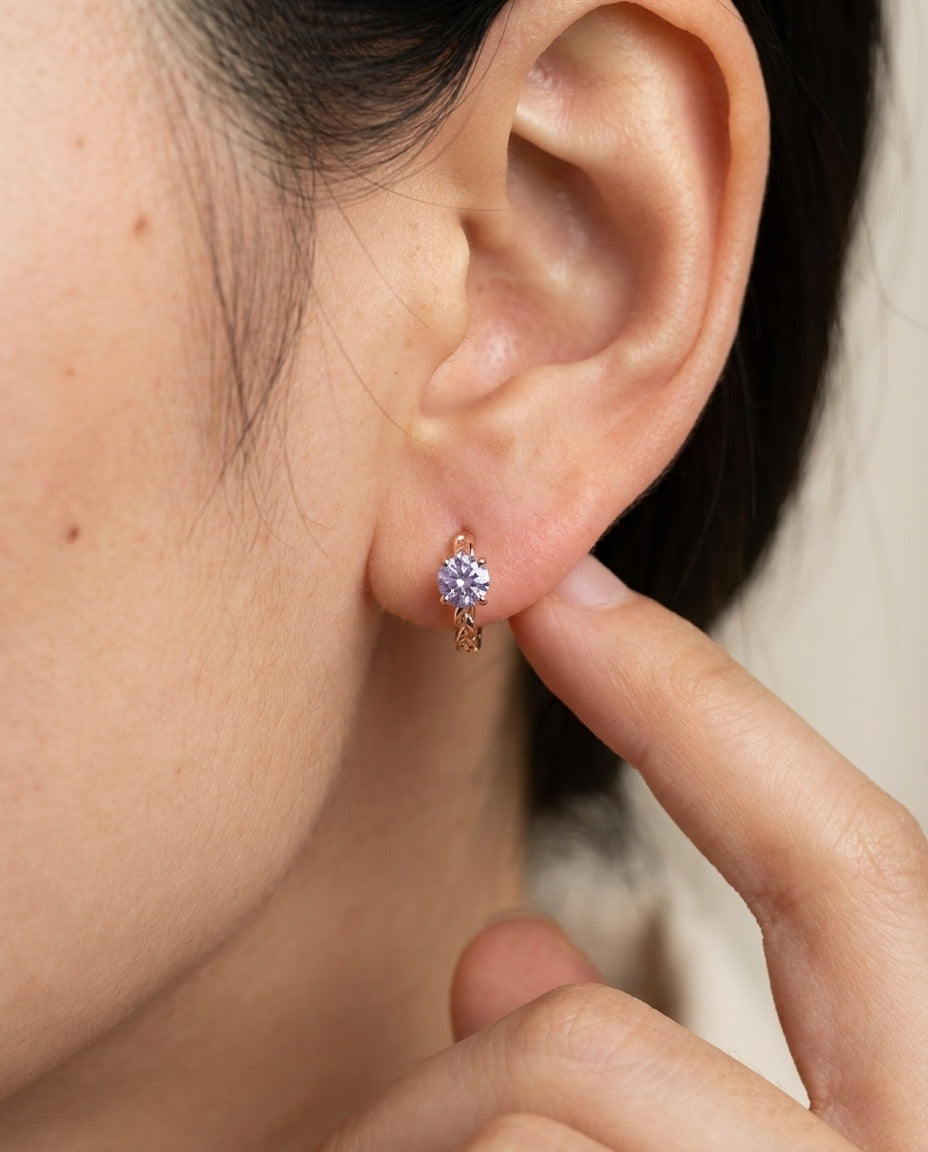 Side profile shot of a model wearing a rose gold vermeil Braided Lavender Moissanite Huggie Hoop (1ctw) with a finger touching the ear.