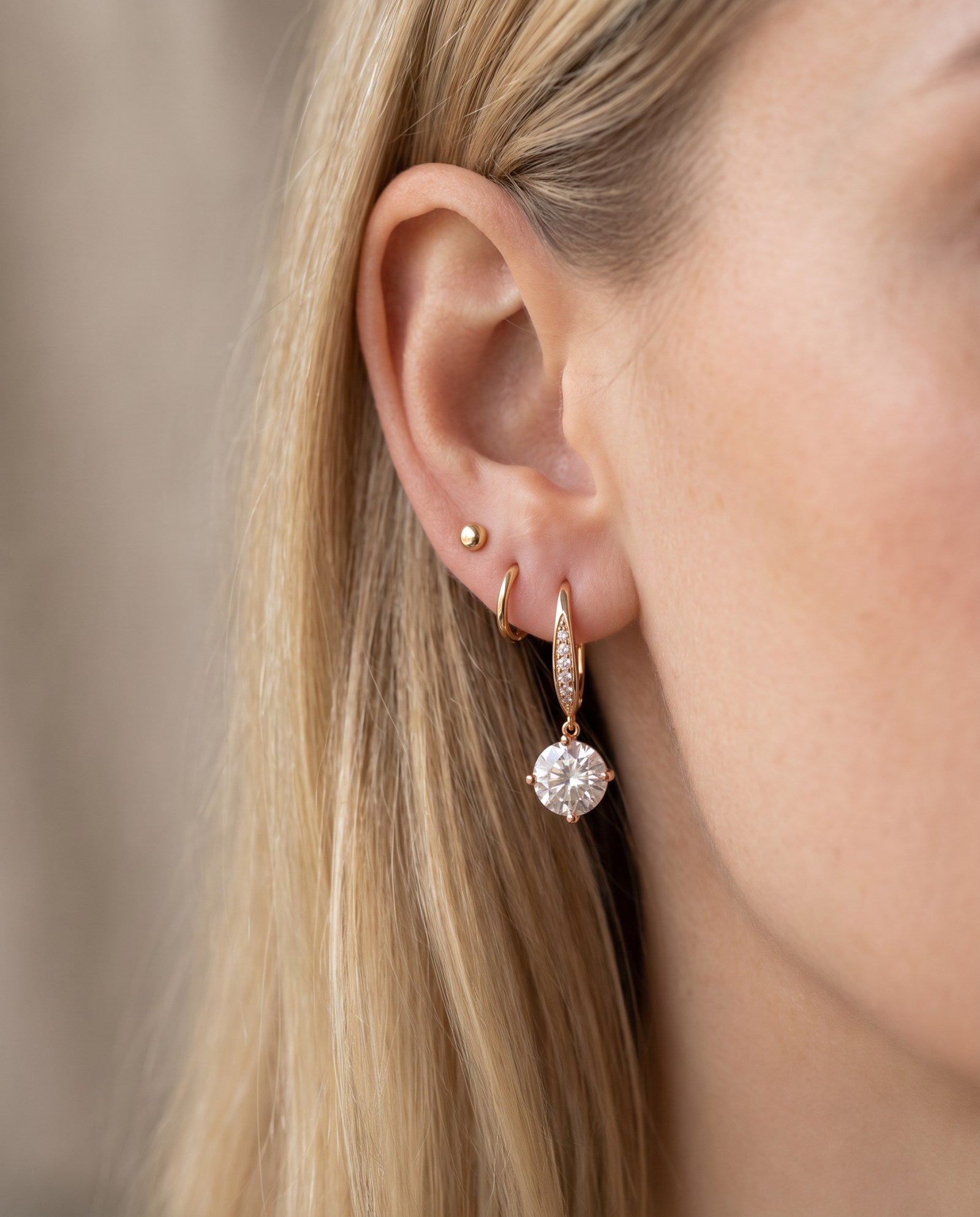 Lifestyle view of a model wearing a rose gold moissanite dangle ear hook stacked alongside small gold huggie hoops.