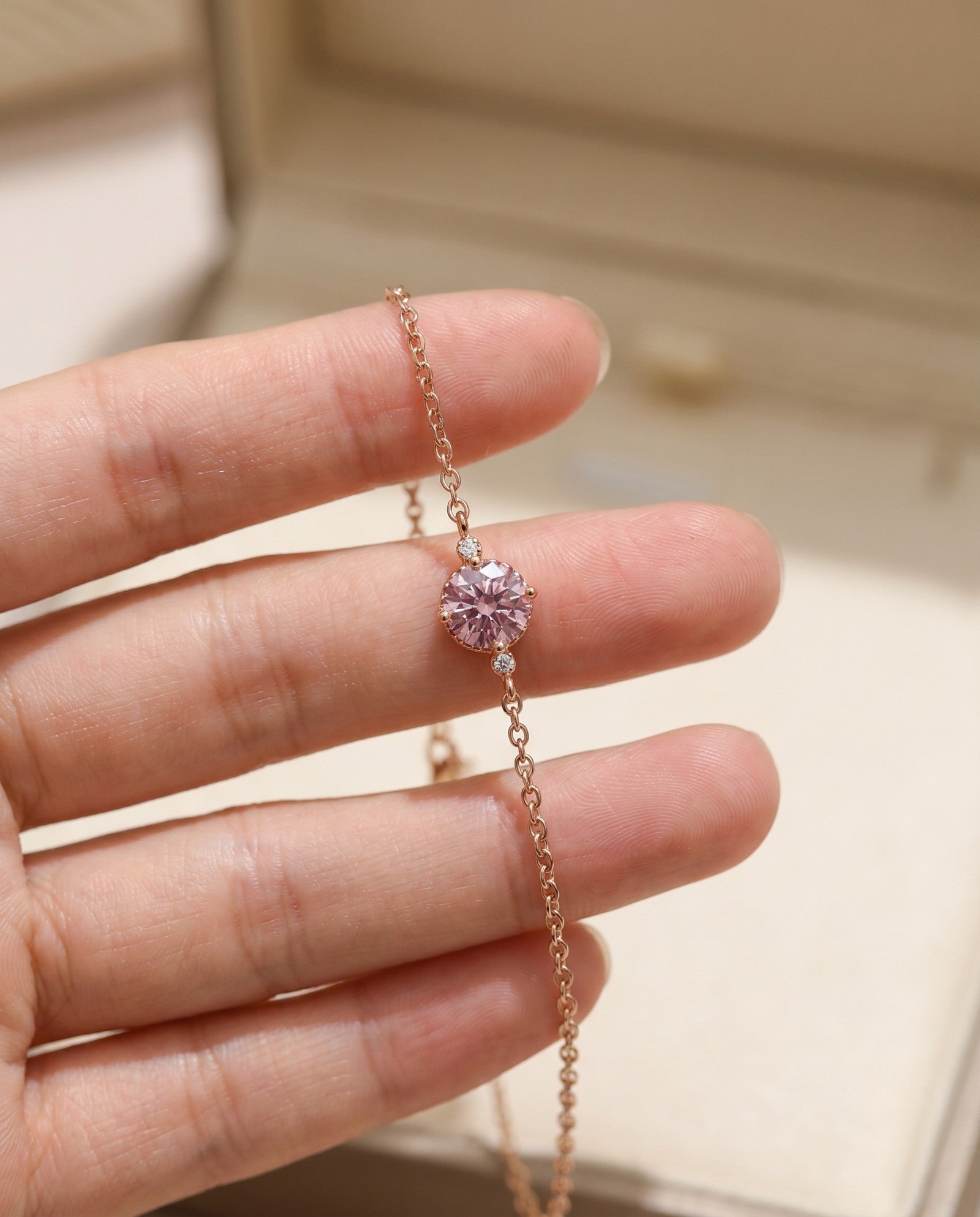 Hand holding a dainty rose gold chain bracelet to showcase the brilliant fire and details of the round 1-carat pink center stone and scalloped basket setting.