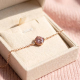 A 18k rose gold vermeil pink moissanite solitaire bracelet resting inside a textured cream-colored linen jewelry gift box under soft sunlight.