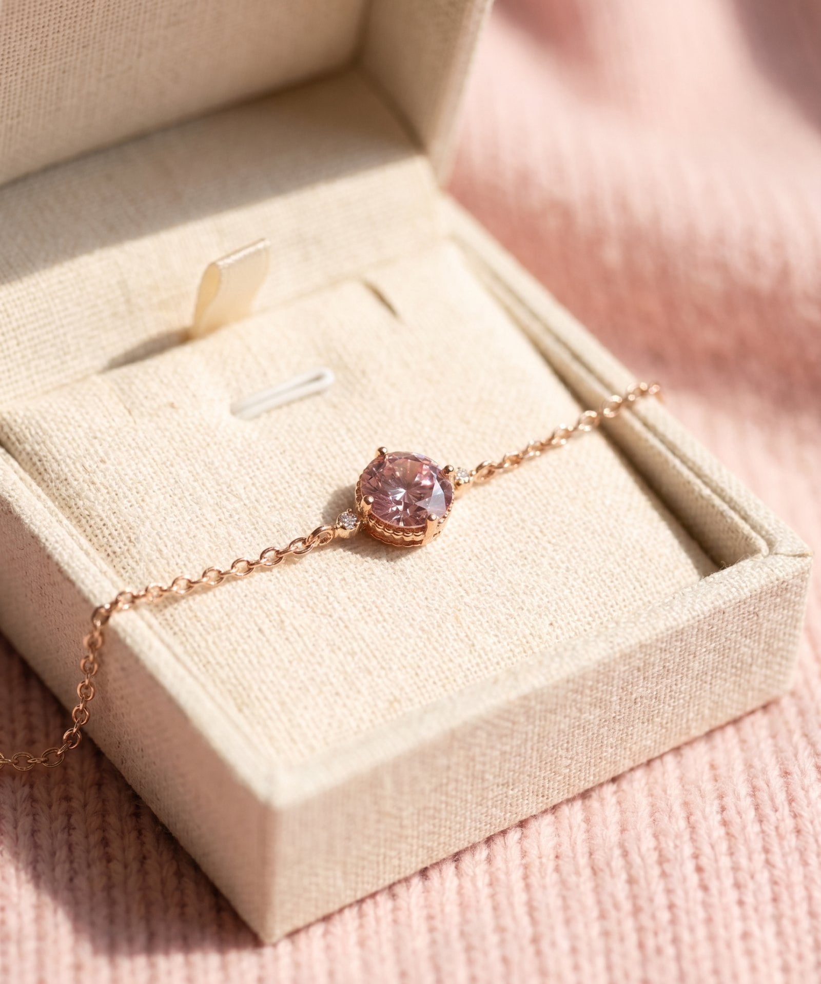 A 18k rose gold vermeil pink moissanite solitaire bracelet resting inside a textured cream-colored linen jewelry gift box under soft sunlight.