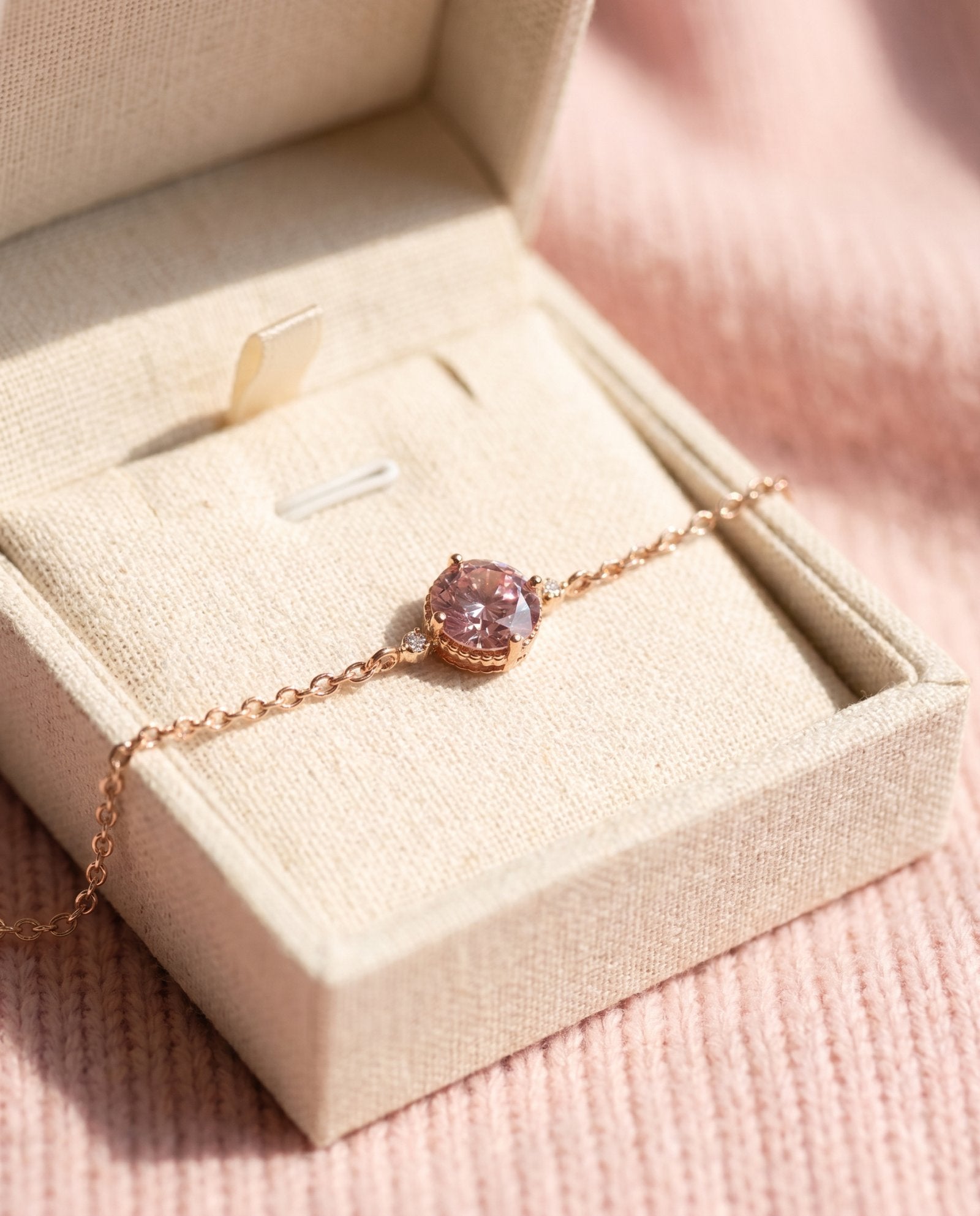 A 18k rose gold vermeil pink moissanite solitaire bracelet resting inside a textured cream-colored linen jewelry gift box under soft sunlight.