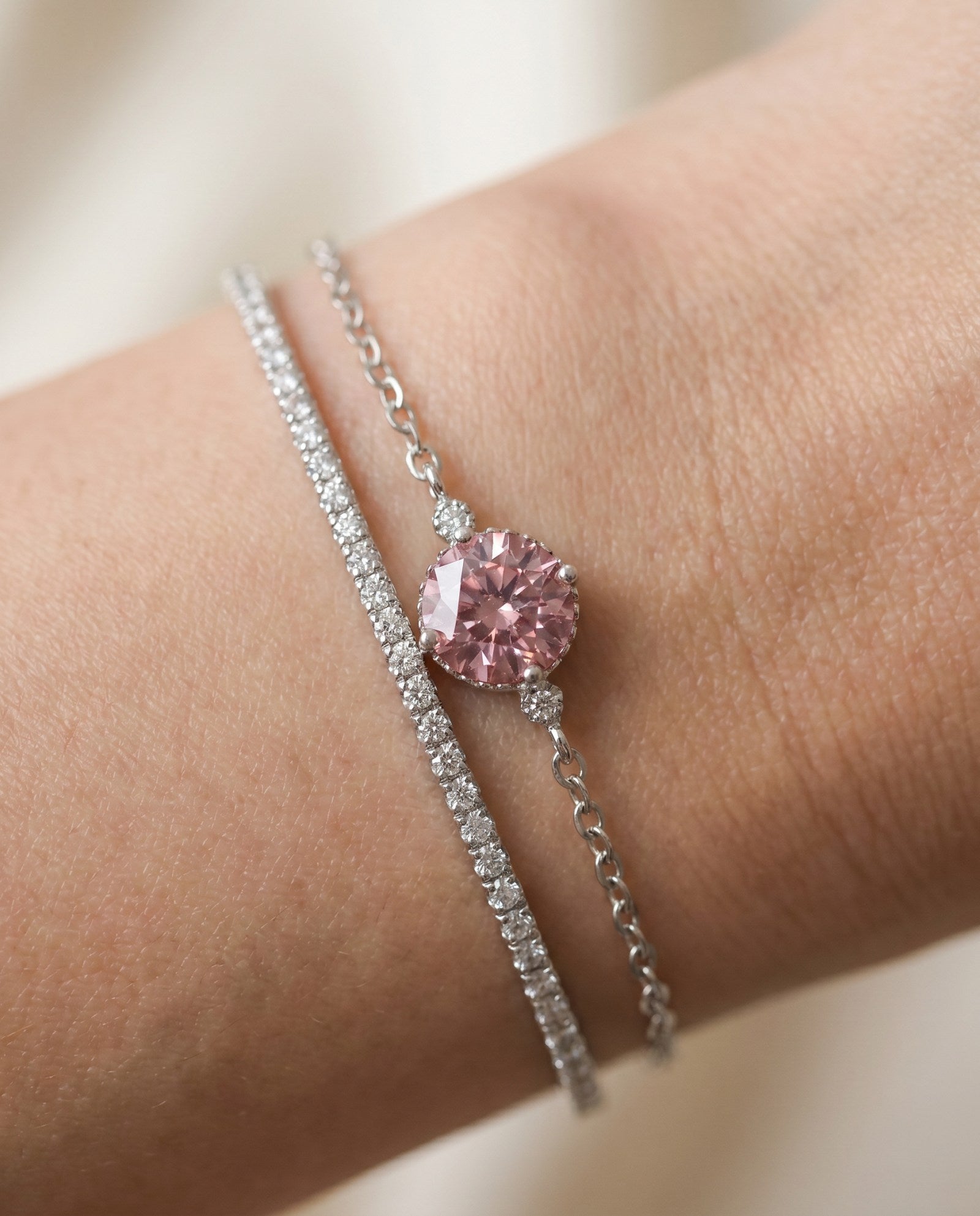 Close-up of a model wearing a dainty silver cable chain bracelet featuring a round-cut rose pink solitaire gemstone, stacked next to a sparkling diamond tennis bracelet.