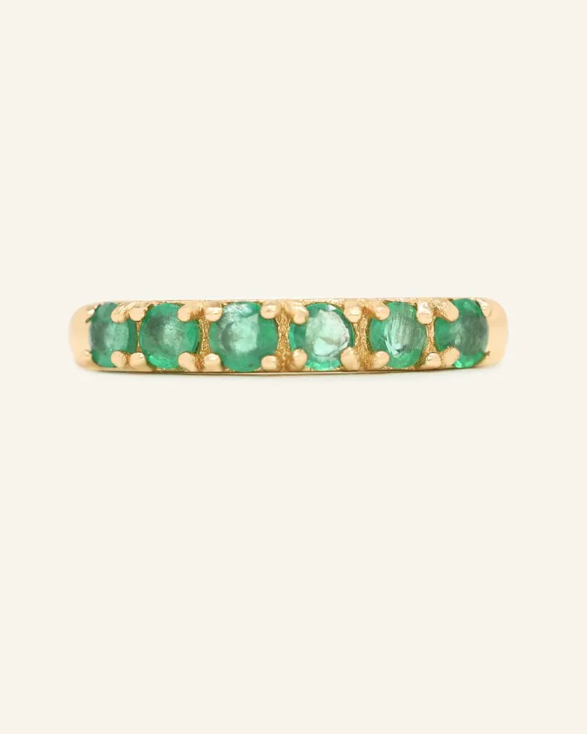 A close-up image of an emerald half eternity band. The ring features a series of vibrant green round-cut emeralds set in a yellow gold band, arranged in a continuous line around half of the band, showcasing a luxurious and elegant design.