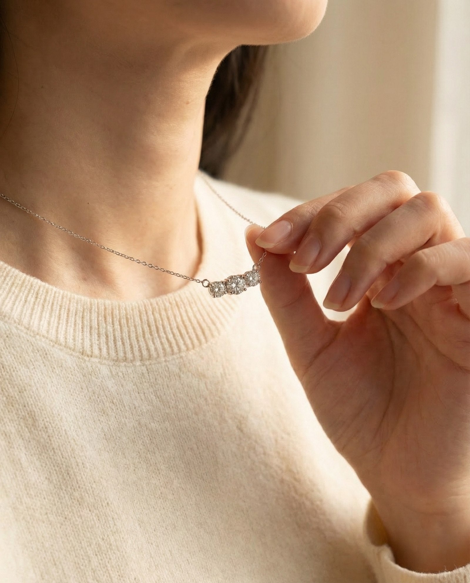 A model wearing the Everglow Trilogy Moissanite Necklace in silver, paired with a cream sweater for a clean, minimalist look.