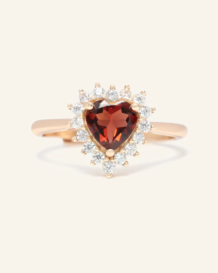A rose gold ring featuring a deep red garnet stone cut in a heart shape, surrounded by a halo of sparkling cubic zirconias, set on a smooth band.
