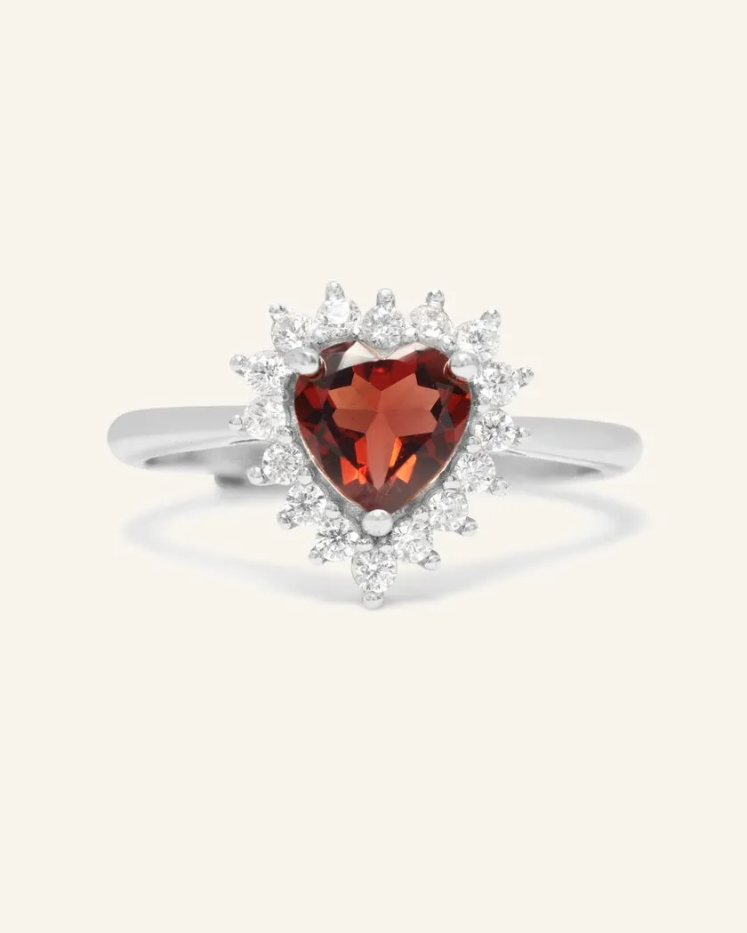 A silver ring featuring a deep red garnet stone cut in a heart shape, surrounded by a halo of sparkling cubic zirconias, set on a smooth band.