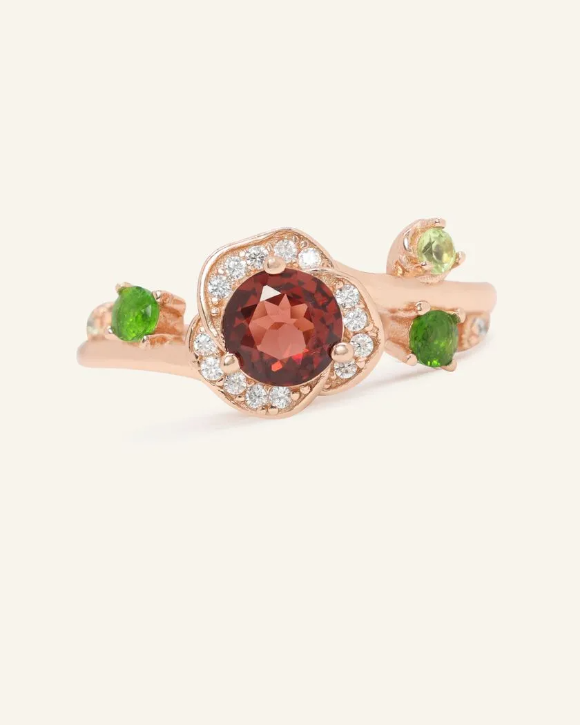 A rose gold ring featuring a central, round-cut garnet stone, surrounded by a halo of cubic zirconias. On either side of the garnet, green diopside gemstones and a light green peridot gemstone are set to mimic leaves in a floral design.