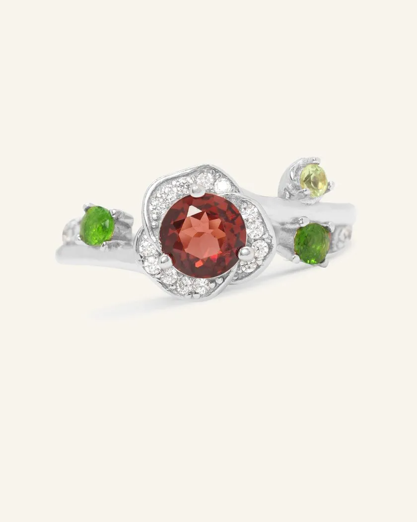 A silver ring featuring a central, round-cut garnet stone, surrounded by a halo of cubic zirconias. On either side of the garnet, green diopside gemstones and a light green peridot gemstone are set to mimic leaves in a floral design.
