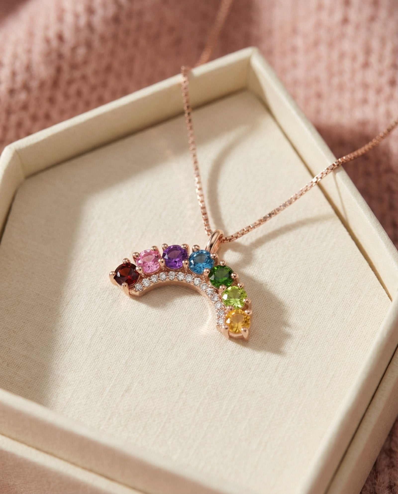 Rose gold rainbow arc necklace featuring garnet, topaz, diopside, and citrine resting in a textured beige jewelry gift box.