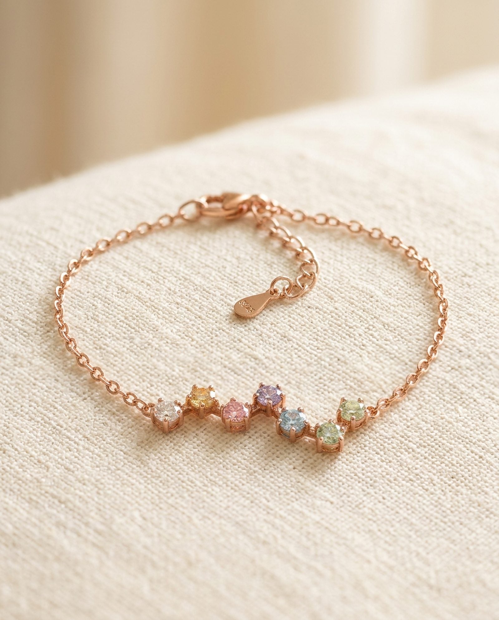 An angled tabletop lifestyle shot of the rose gold Pastel Constellation Moissanite Bracelet, showcasing the fine chain, clasp detail, and the wave-like constellation pattern of the multi-colored gems.
