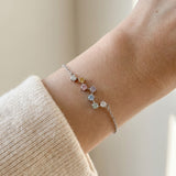 A close-up lifestyle photograph of the silver Pastel Constellation Moissanite Bracelet on a model's wrist, showing the subtle flash of the multi-colored pastel stones against a cream-colored sweater.