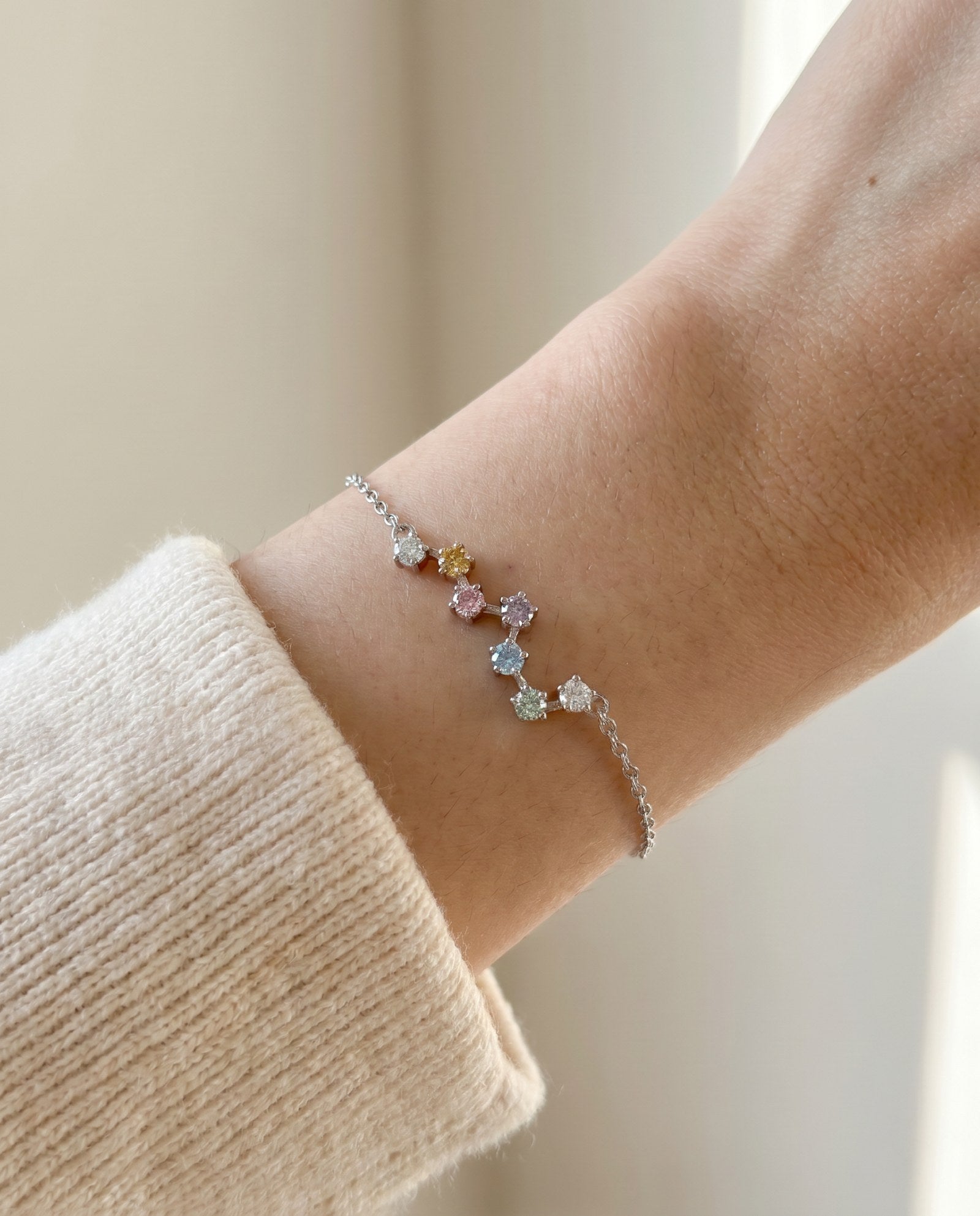 A close-up lifestyle photograph of the silver Pastel Constellation Moissanite Bracelet on a model's wrist, showing the subtle flash of the multi-colored pastel stones against a cream-colored sweater.