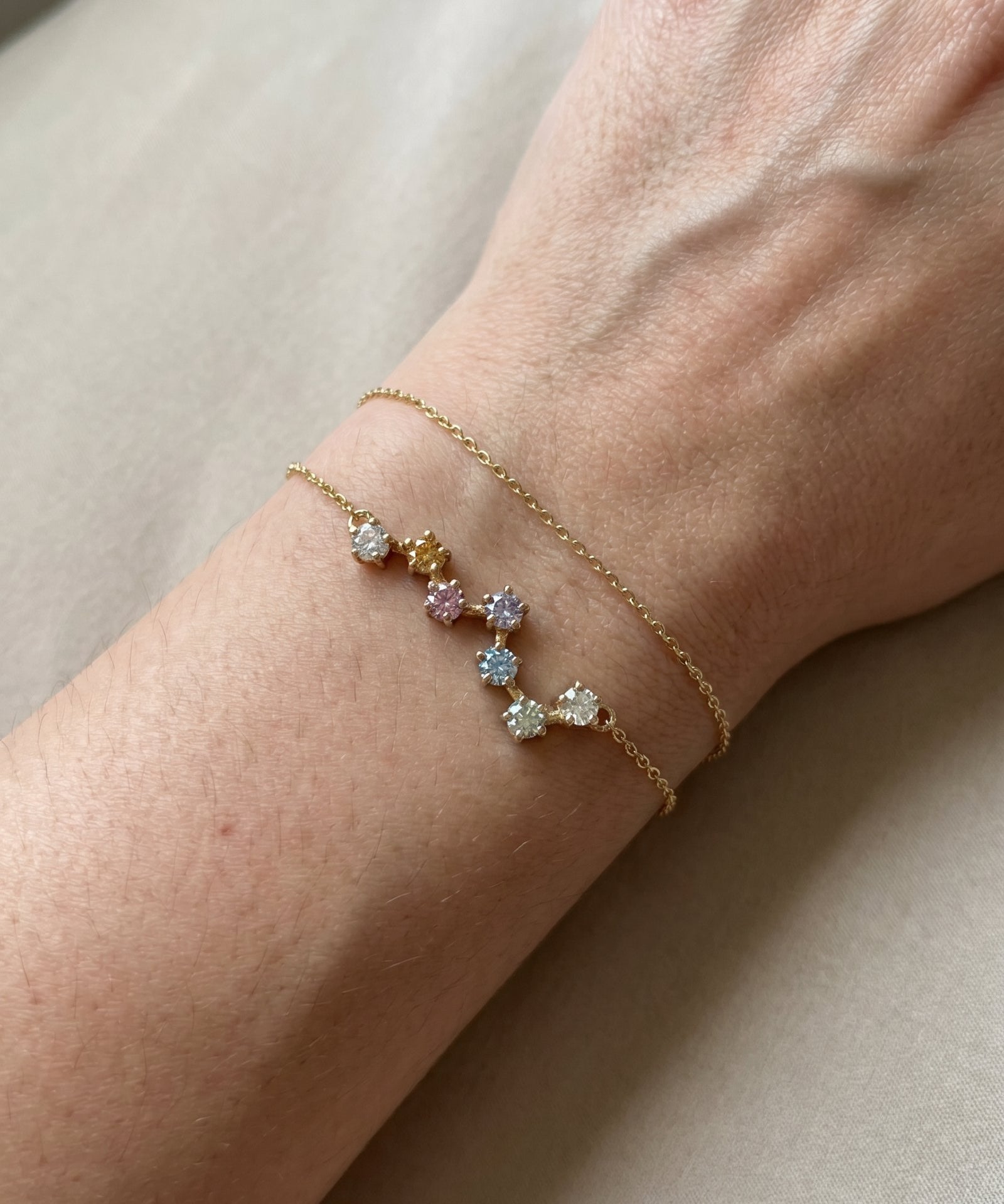 A close-up lifestyle photograph of the yellow gold Pastel Constellation Moissanite Bracelet being worn on a woman's wrist alongside a thin matching gold chain, demonstrating its delicate scale.