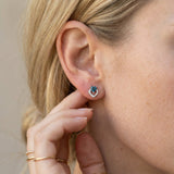 Lifestyle photograph of a model wearing teal-blue tourmaline heart earrings, styled with minimal gold rings.
