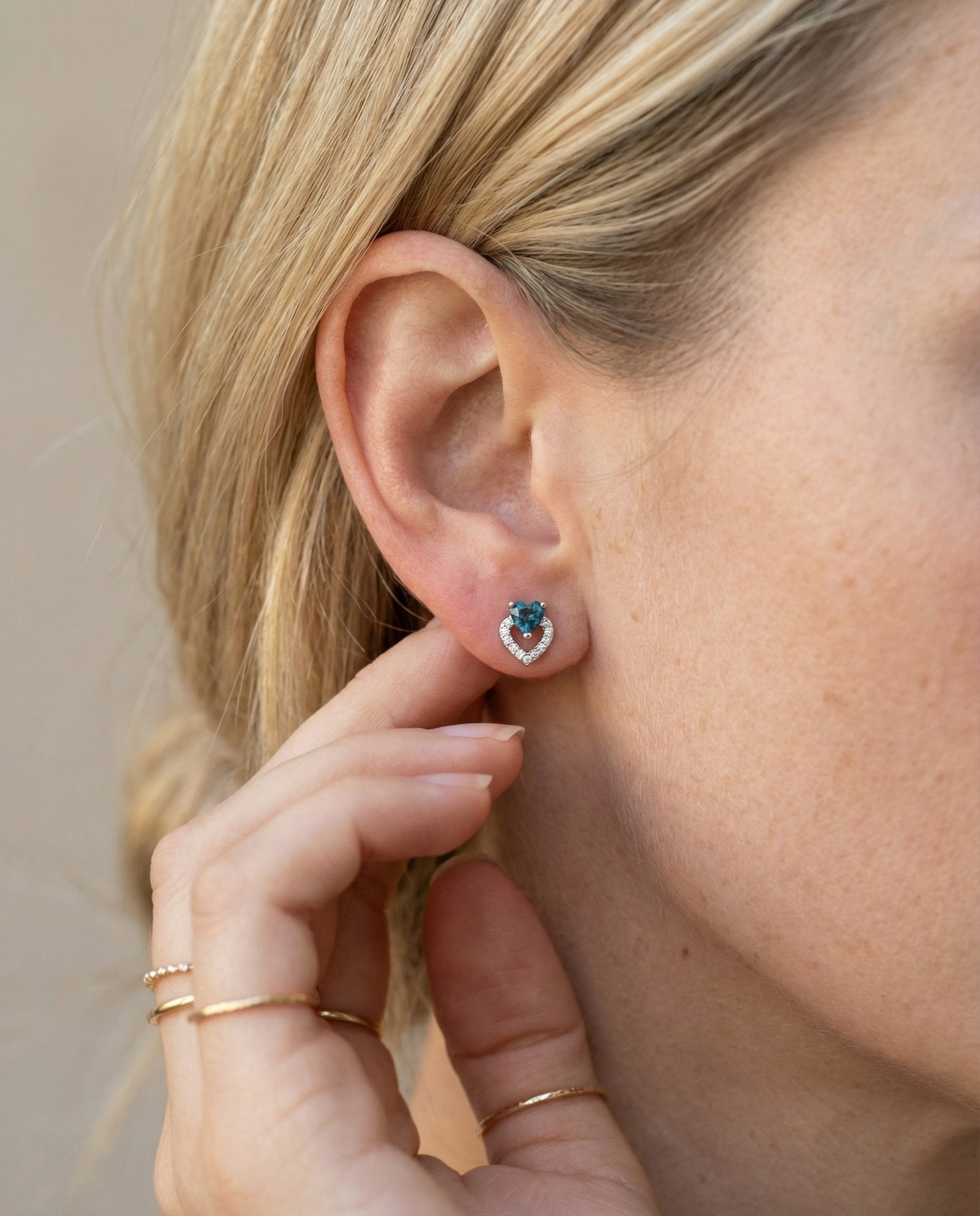 Lifestyle photograph of a model wearing teal-blue tourmaline heart earrings, styled with minimal gold rings.