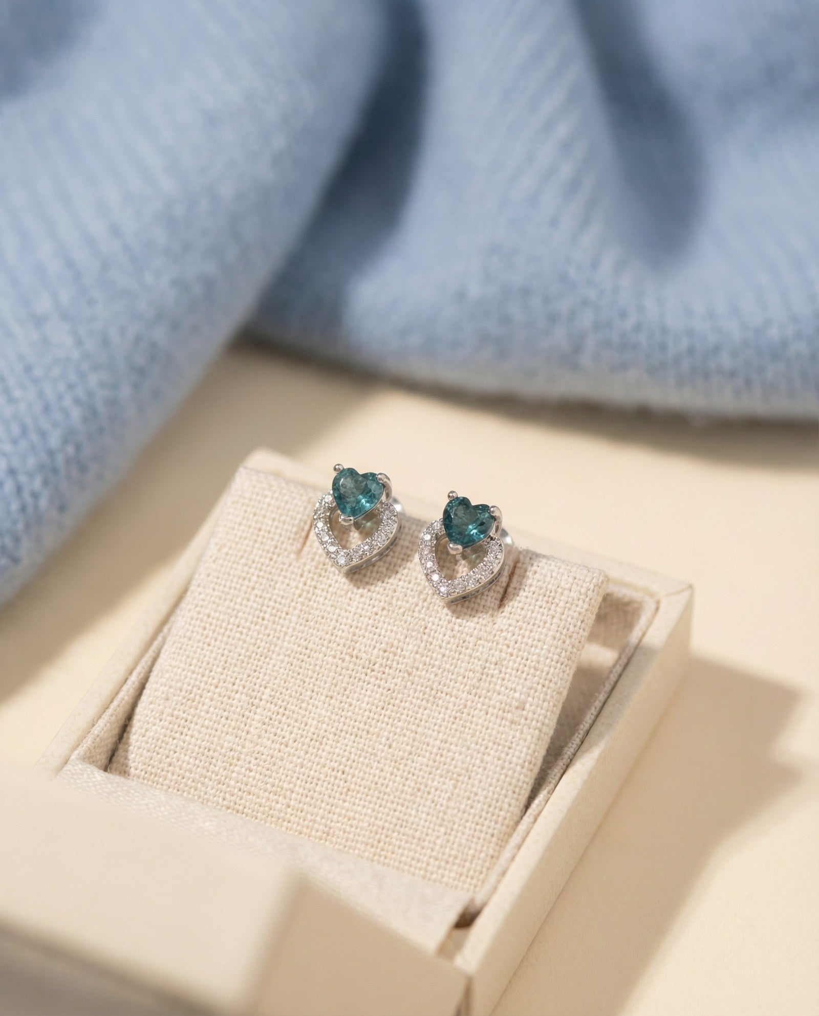Close-up of Petite Blue Tourmaline Heart Studs in a cream jewelry box with a soft blue knit background.