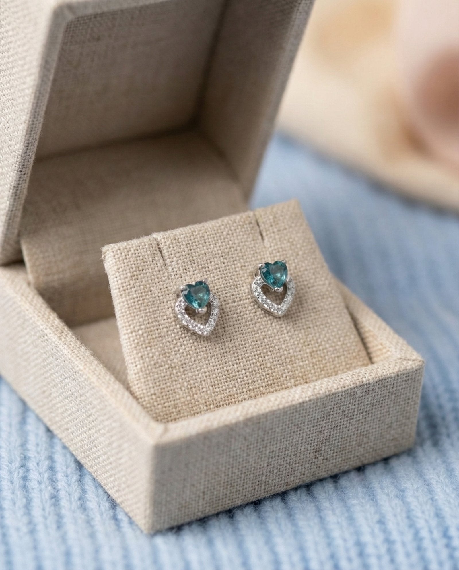 Angled view of heart-cut blue tourmaline earrings with a shimmering pavé heart frame in a luxury gift box.