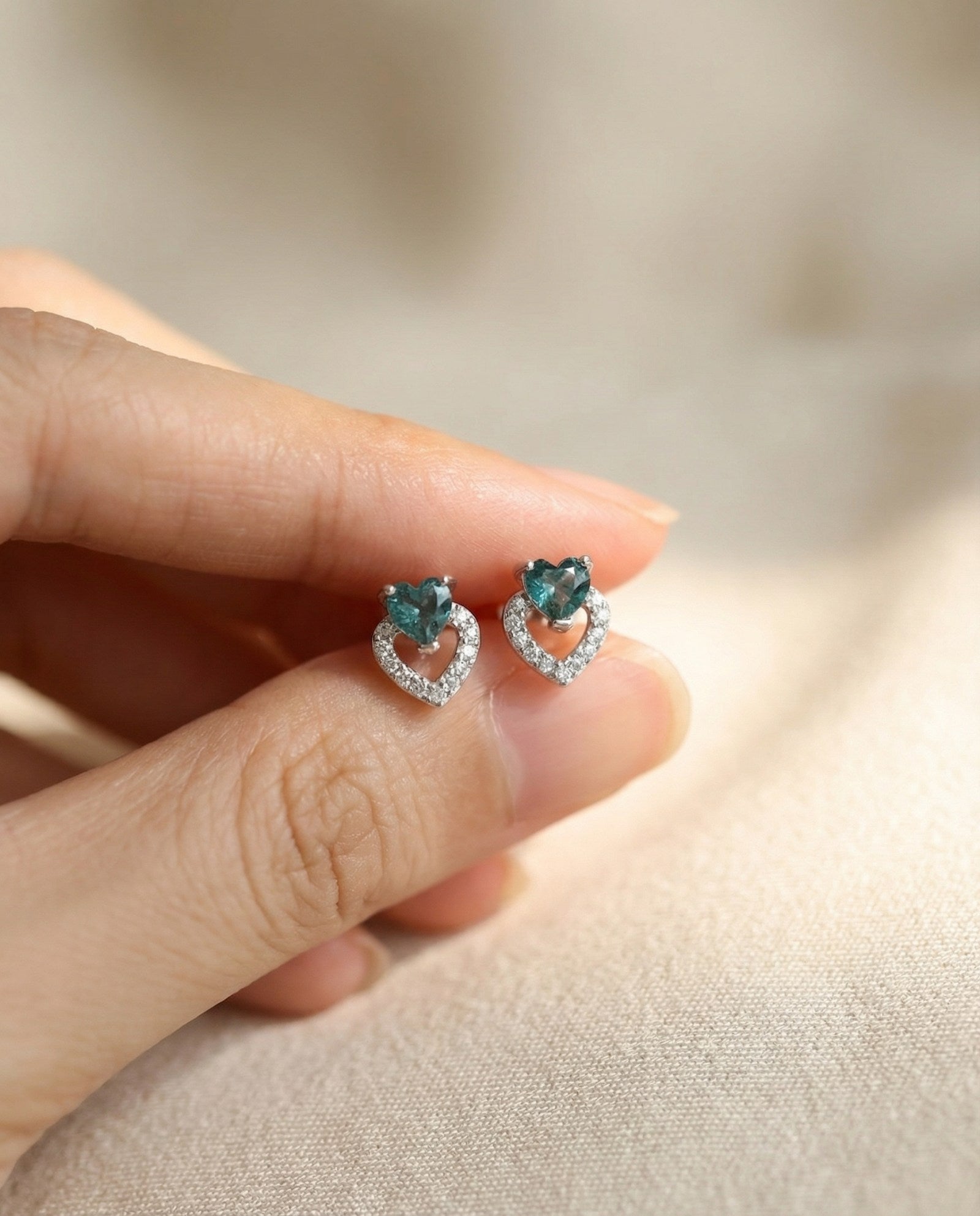 Hand-held view of Petite Blue Tourmaline Heart Studs in silver to show scale and the vibrant blue-green gemstone color.