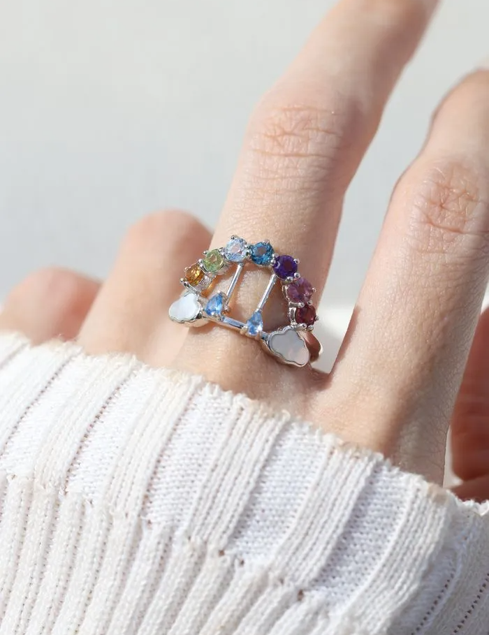 Rainbow's Promise Ring worn on a finger, showcasing a gradient of citrine, peridot, topaz, amethyst, tourmaline, and rhodolite gemstones, accented with delicate cloud motifs and movable raindrop details.