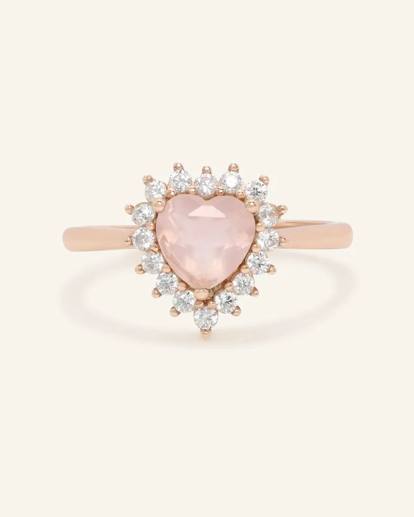 A rose gold ring featuring a heart-shaped rose quartz center stone, surrounded by a halo of sparkling cubic zirconias.