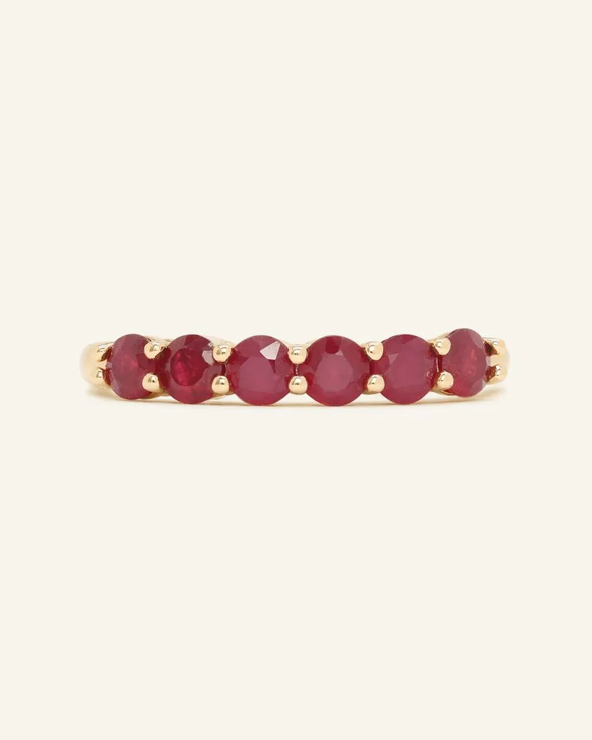 A close-up image of a ruby half eternity band. The ring features a series of vibrant, deep red round-cut rubies set in a yellow gold band, arranged in a continuous line around half of the band, showcasing a timeless and elegant design.