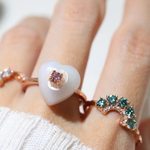 A model wearing the delicate Sakura Jadeite Heart Ring in rose gold, featuring a jadeite heart centerpiece, a sparkling pink moissanite as the focal stone, and intricately carved mother-of-pearl flower petals for a touch of elegance.