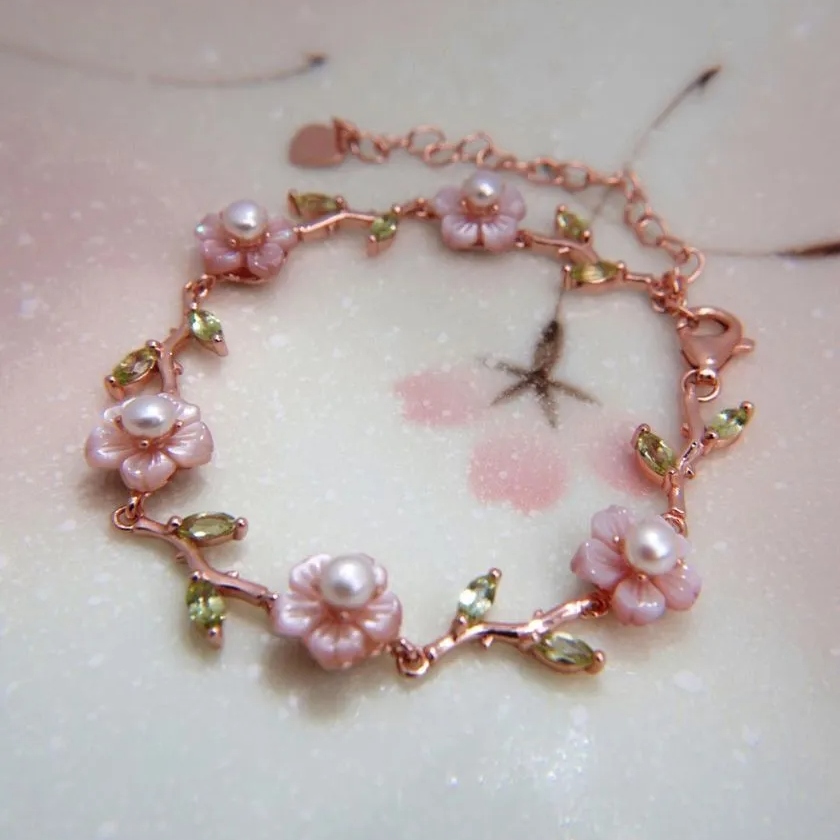 A delicate sakura twig bracelet featuring hand-carved mother-of-pearl flowers with freshwater pearl centers and peridot gemstone leaves, set in a rose gold finish. Elegant and nature-inspired.