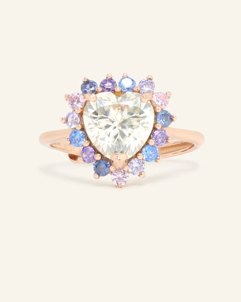 A close-up image of a ring featuring a heart-shaped white moissanite center stone surrounded by a halo of smaller round cubic zirconias in shades of blue, purple, and pink, set in a rose gold band.