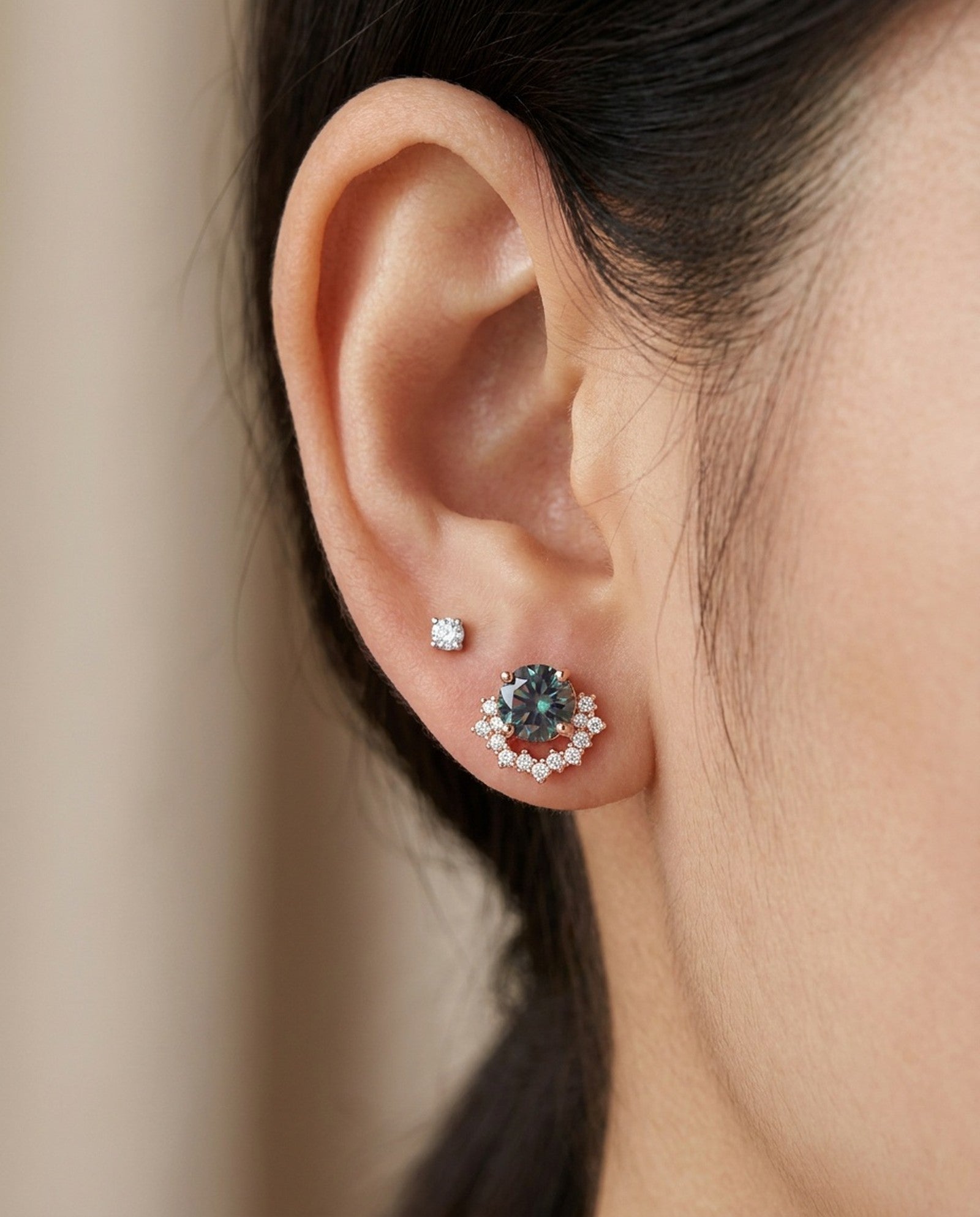 Model wearing 18k rose gold Starlight Crown Crescent Moissanite Stud in Celestial Teal in lower ear lobe piercing next to a clear solitaire stud.