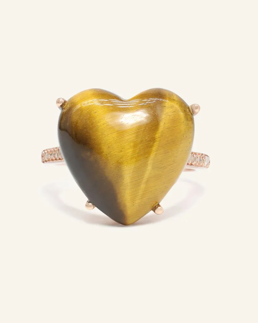 A close-up image of a tiger eye heart ring. The ring features a large, polished heart-shaped tiger eye gemstone set in a rose gold band, with the stone's characteristic golden-brown hues and chatoyancy creating a striking visual effect.