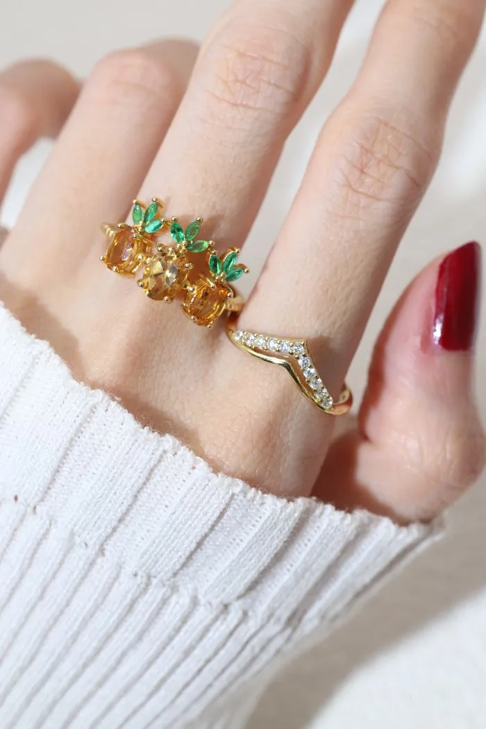 A model wearing a trio of pineapple-themed rings featuring yellow citrine gemstones mimicking the fruit's body, with green cubic zirconia leaves at the top, all set in a yellow gold band.