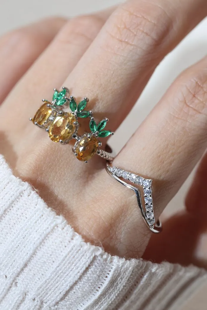 A model wearing a trio of pineapple-themed rings featuring yellow citrine gemstones mimicking the fruit's body, with green cubic zirconia leaves at the top, all set in a silver band.