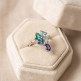 Silver blue topaz clover ring with dangling violet iolite displayed on an ivory jewellery box