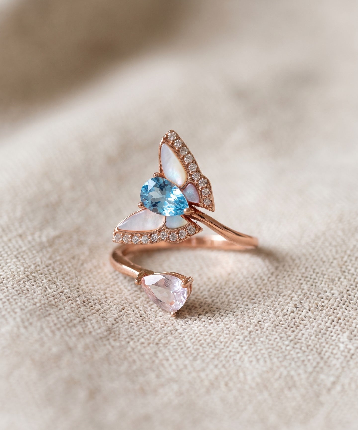 Rose gold butterfly ring with Swiss blue topaz, rose quartz and mother-of-pearl wings resting on textured ivory linen