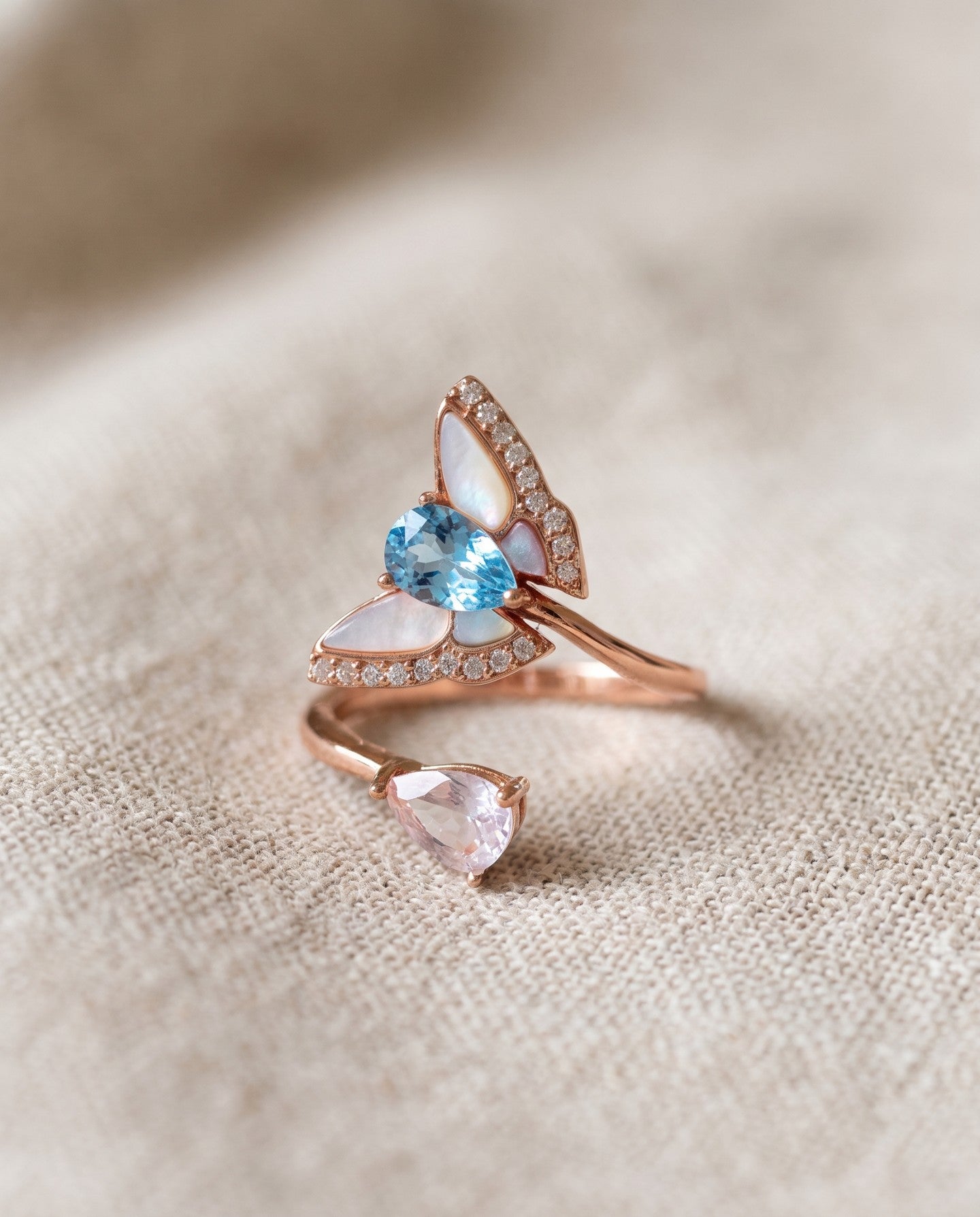 Rose gold butterfly ring with Swiss blue topaz, rose quartz and mother-of-pearl wings resting on textured ivory linen